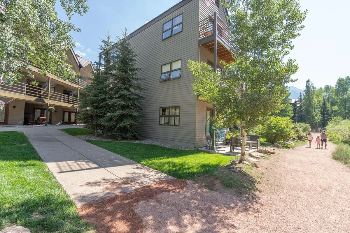 A tree-lined pathway leads to a modern building facade with balconies featuring railings. The scene is lively, with people strolling, enjoying the outdoor space, and socializing. The combination of building and nature highlights both accessibility and a sense of community.