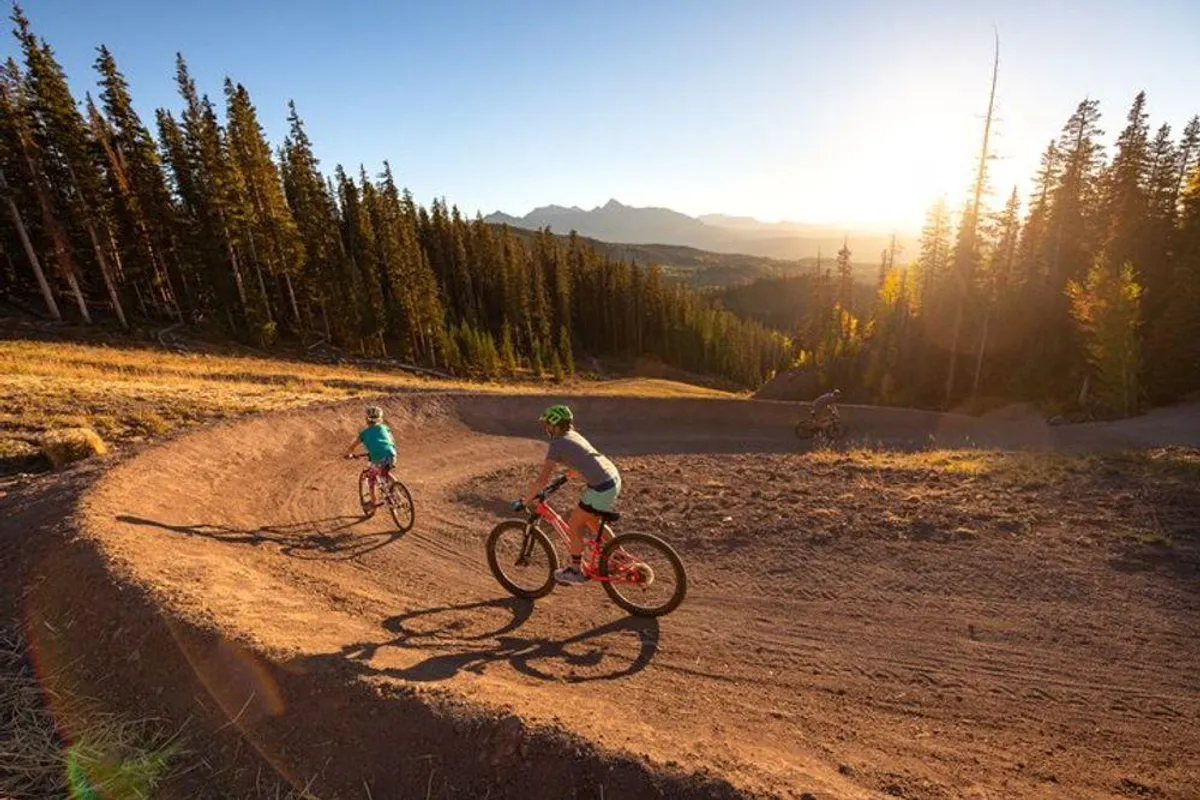 A curving bike path winds through rugged mountainous terrain, with bicyclists actively traversing the trail. The backdrop of dramatic peaks and lush greenery highlights Telluride’s appeal for mountain biking, hiking, and outdoor summer adventures.