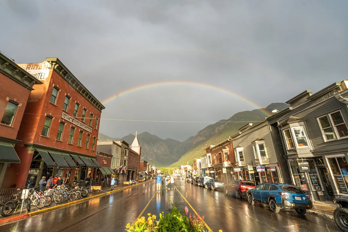 This image captures Telluride's vibrant historic downtown with charming buildings, a rainbow arching over wet streets, and people walking and biking. The energy of the street reflects the lively, walkable character of the town and the variety of shops and cafes easily accessible by foot.