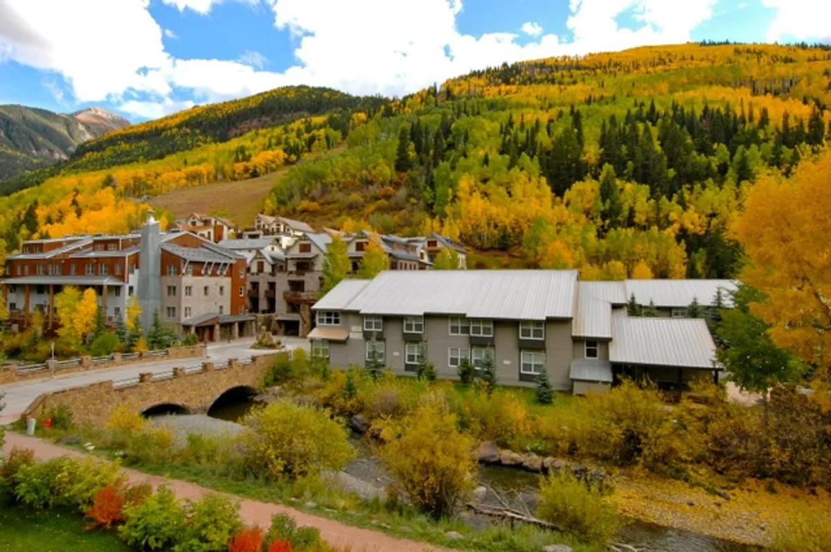 A scenic riverside with colorful autumn foliage, a bridge, and nearby buildings illustrates the blend of nature and community in Telluride. The river and pathways invite residents and visitors to explore and relax outdoors.