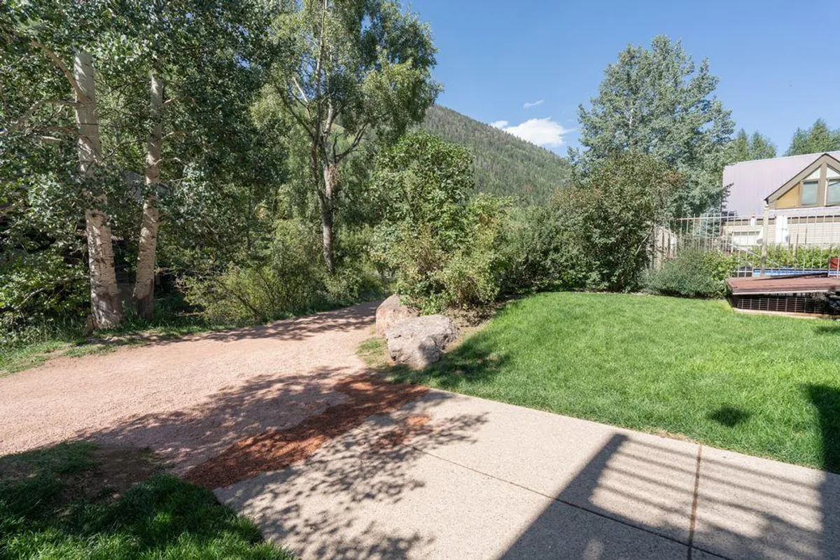 A scenic grassy area bordered by a winding pathway, surrounded by lush trees and rock formations with distant mountain views. The space is open and welcoming, ideal for outdoor activities such as walking, picnicking, or simply relaxing in nature.
