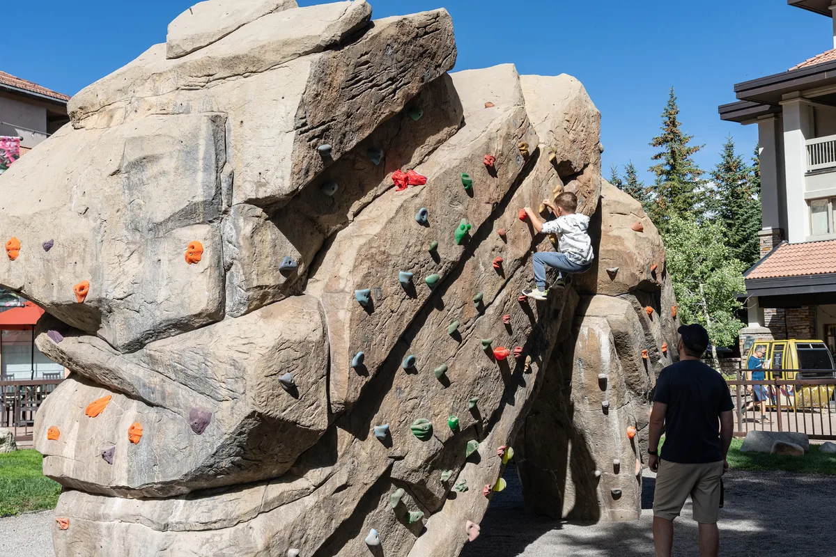 A vibrant outdoor climbing rock structure catches the eye, with a child actively climbing and an adult nearby offering encouragement and support. The scene radiates energy and the joy of family bonding through playful exploration.