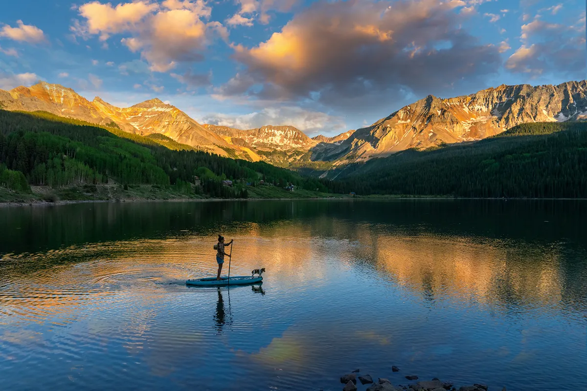 A paddleboarder, accompanied by a dog, glides across calm water with mountains reflected in the surface behind. The tranquil setting captures leisurely river adventures and the peaceful, scenic aspect of Telluride’s outdoor lifestyle.