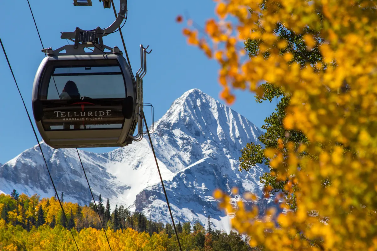 The image features the Telluride gondola lift ascending towards snow-capped mountains, with vivid autumn foliage below. It emphasizes both easy access to outdoor adventure and the town's stunning natural setting.