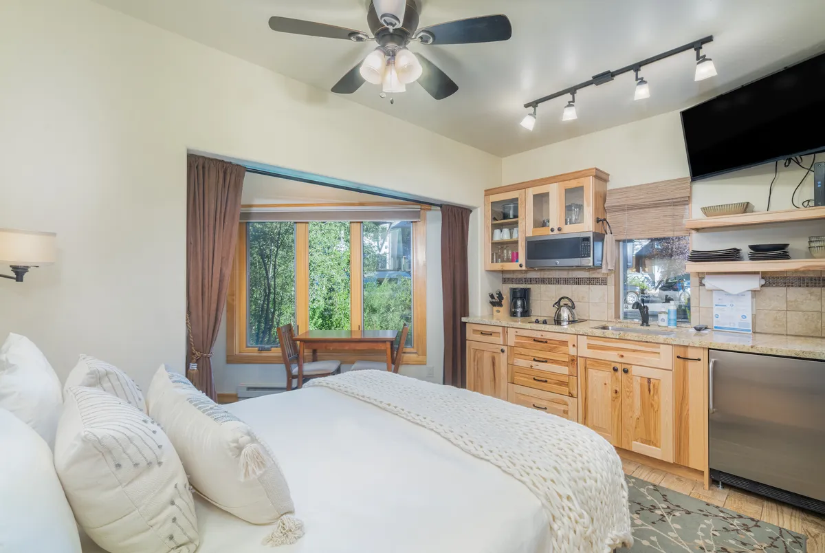 The image showcases a cozy studio interior with a comfortable bed adorned with decorative pillows, a well-equipped kitchenette, and a small dining table positioned by a window that lets in natural light. The overall atmosphere is warm and inviting, ideal for relaxing, cooking, or enjoying a meal with a view.