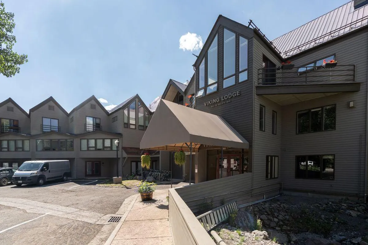 The welcoming entrance of the Viking Lodge is shown, featuring the lodge’s sign, main entrance awning, a bicycle rack, and the architectural exterior. The scene captures a vibrant, social atmosphere with guests arriving or gathering outside.