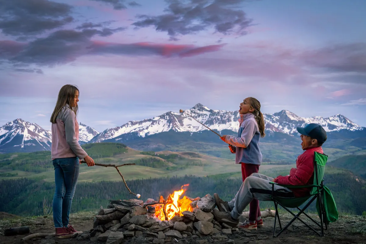 Children gathered around a campfire, toasting marshmallows under the watchful eye of an adult, with mountains forming a stunning backdrop. The mood is joyful, blending the excitement of outdoor activity with the warmth of family togetherness.