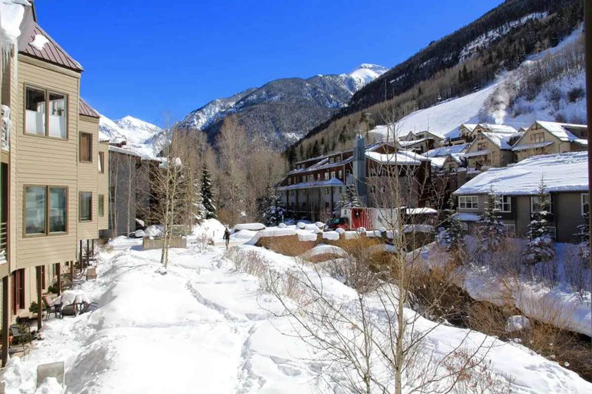 A serene winter scene with snow-covered ground, mountain backdrop, and a pathway leading through the property. The apartment buildings are visible, highlighting the property’s location in a mountainous, alpine environment. The image evokes Telluride’s seasonal climate and winter recreational appeal.