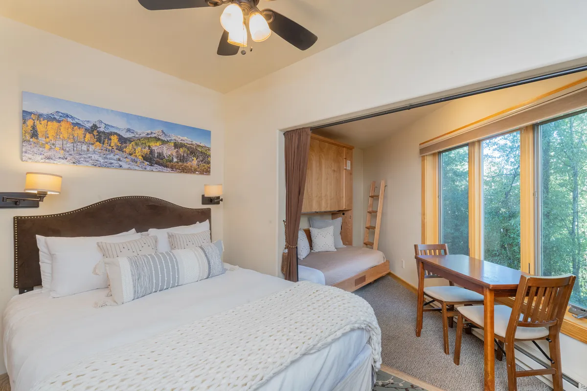A sun-filled sleeping and dining area is shown, with a large bed adorned with decorative pillows, a dining table and chairs, prominent artwork of a mountain landscape, and generous natural light streaming through the windows. The mood is both warm and inviting, highlighting the bright, relaxing vibe of the studio.