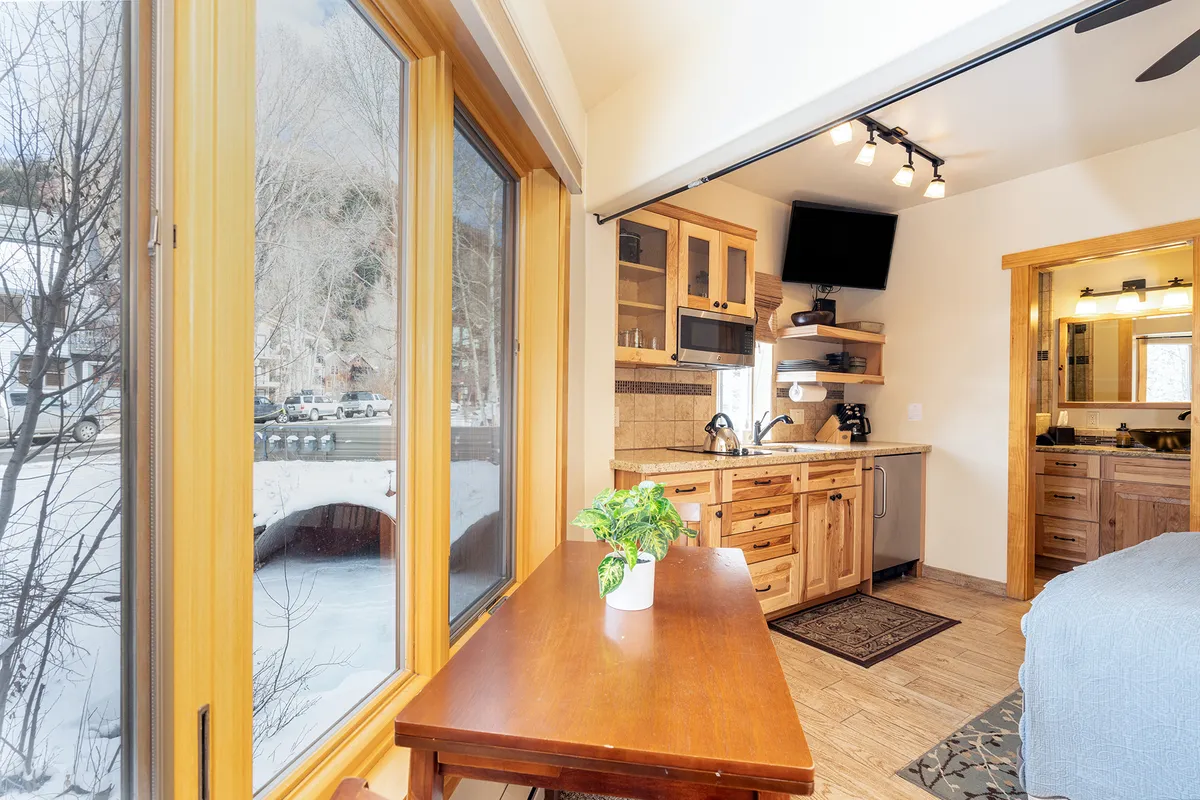 Featuring a wooden table, kitchen cabinetry, and large windows that let in natural light, this image conveys the atmosphere of a comfortable space for meal preparation or family dining. The setting feels homey and practical, with enough room for multiple activities, reinforcing the sense of ease and accessibility.
